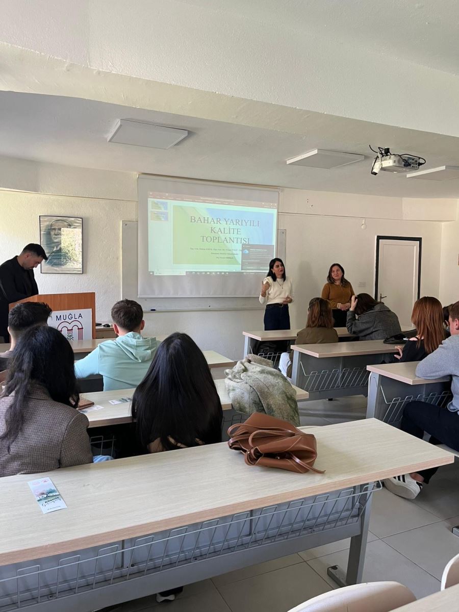 Bir üniversite sınıfında gerçekleştirilen kalite toplantısında öğrenciler sıralarda otururken iki öğretim elemanı sınıfın önünde sunum yapmaktadır. Projektör ekranında “Bahar Yarıyılı Kalite Toplantısı” başlıklı bir sunum yer almaktadır. Öğrenciler dikkatle sunumu dinlemektedir. Sınıfın ön kısmında bir kürsü ve Muğla Sıtkı Koçman Üniversitesi logosu görülmektedir. Sunumu gerçekleştiren öğretim elemanları öğrencilerle iletişim kurarak toplantıyı yürütmektedir.